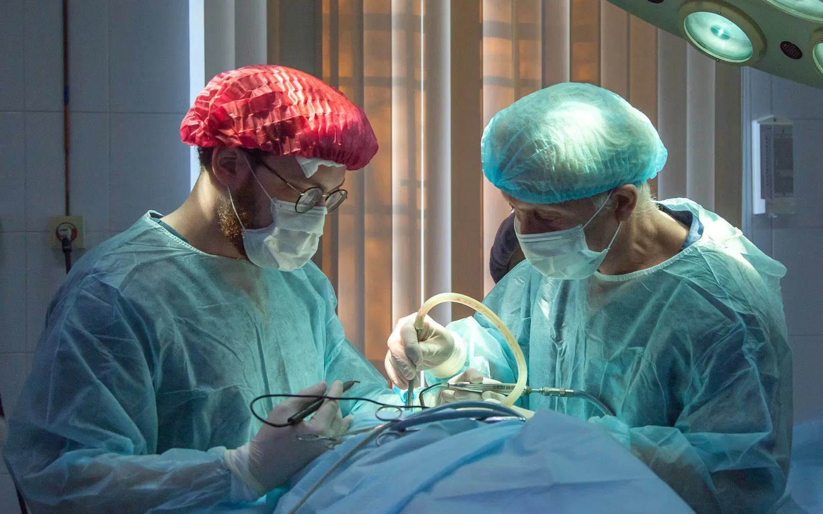 Two surgeons wearing surgical gowns, masks, and caps performing an operation under bright lights.