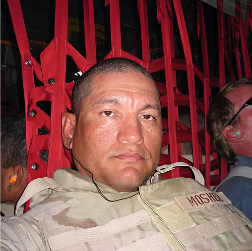 Man in military uniform with name tag "MOSTRAN" seated against red cargo netting inside aircraft.