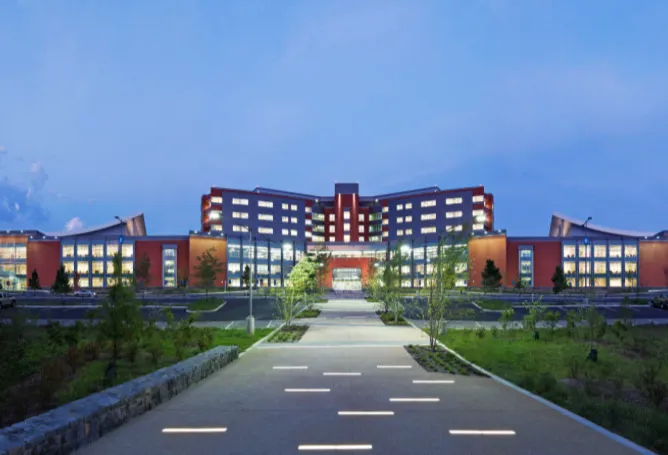 Modern multi-story building with extensive glass windows, illuminated at dusk, and surrounding greenery.
