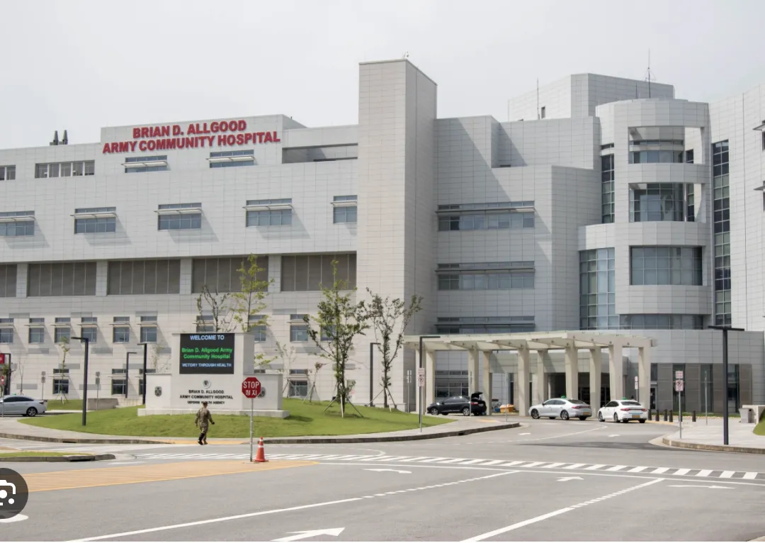 Exterior view of Brian D. Allgood Army Community Hospital with vehicles and a pedestrian near the entrance.