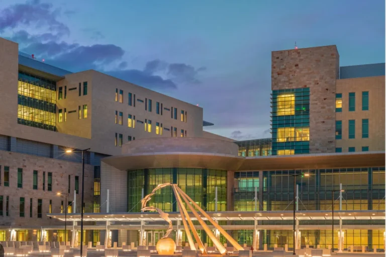 Modern multi-story building with illuminated windows and a large abstract sculpture in front during dusk.