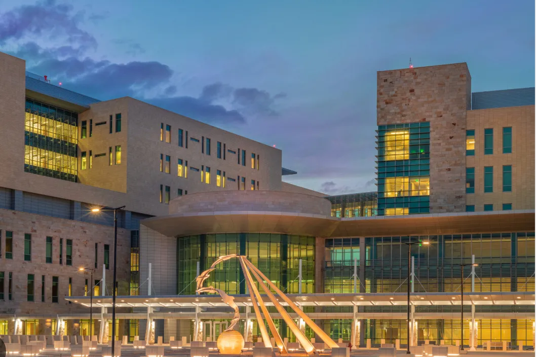 Modern multi-story building with illuminated windows and a large abstract sculpture in front during dusk.