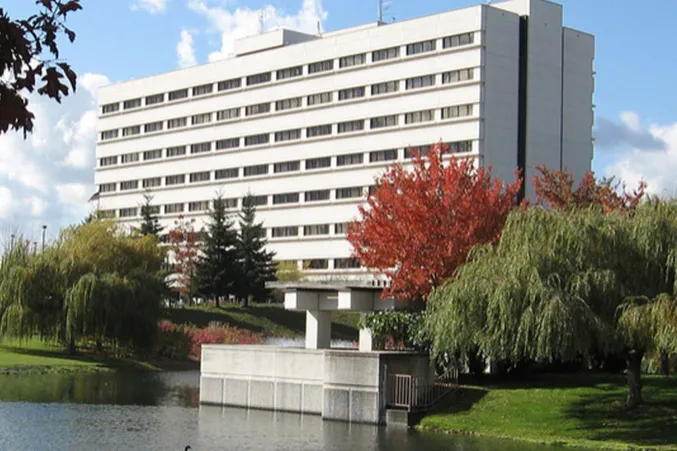 Large white multi-story building behind a pond with autumn trees and greenery.