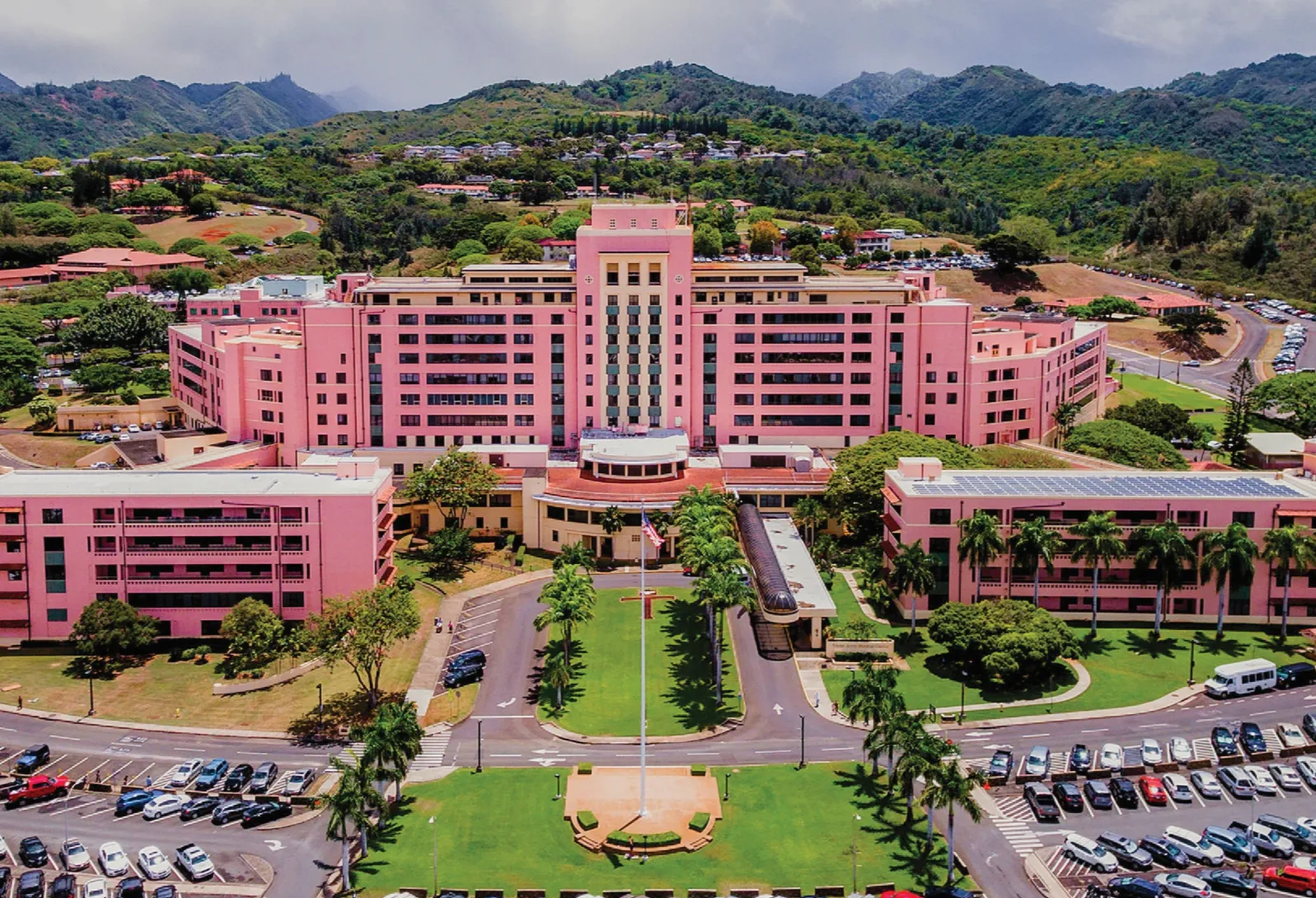 Aerial view of a large pink hospital complex with surrounding parking lots and green mountainous landscape.
