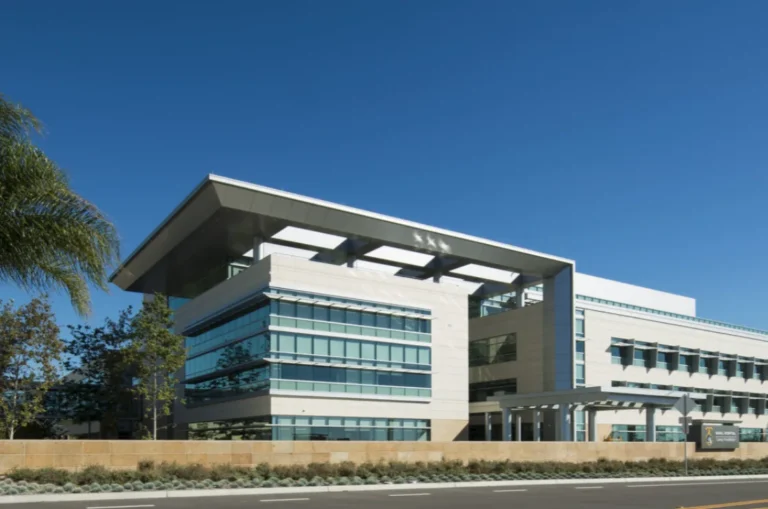 Modern multi-story office building with large glass windows and a flat overhanging roof under clear blue sky.