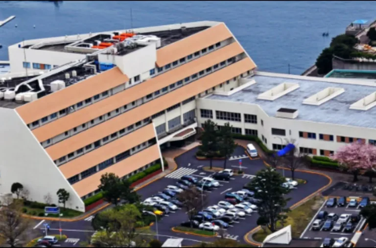 Aerial view of a large, multi-story building with a beige roof and adjacent parking lot near a body of water.
