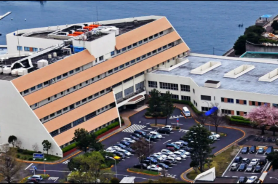Aerial view of a large, multi-story building with a beige roof and adjacent parking lot near a body of water.