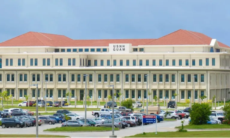 Large beige institutional building with red roof labeled "USNH Guam," surrounded by parked cars.