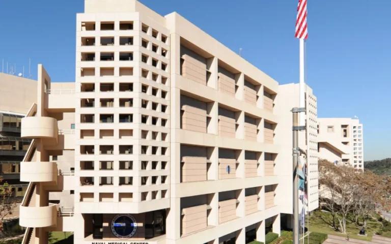 Modern beige multi-story naval medical center building with American flag on clear day.