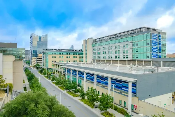 Modern hospital complex with multi-story buildings, green trees lining the street, and a clear blue sky.