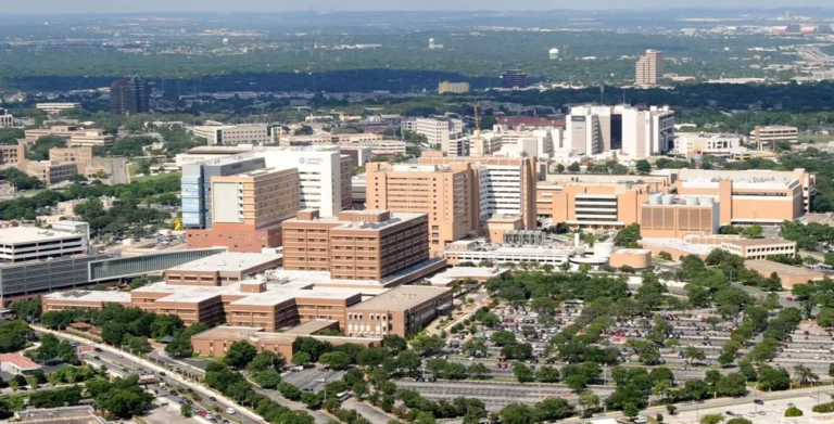 Aerial view of a large hospital complex with multiple beige and white buildings surrounded by trees and parking lots.