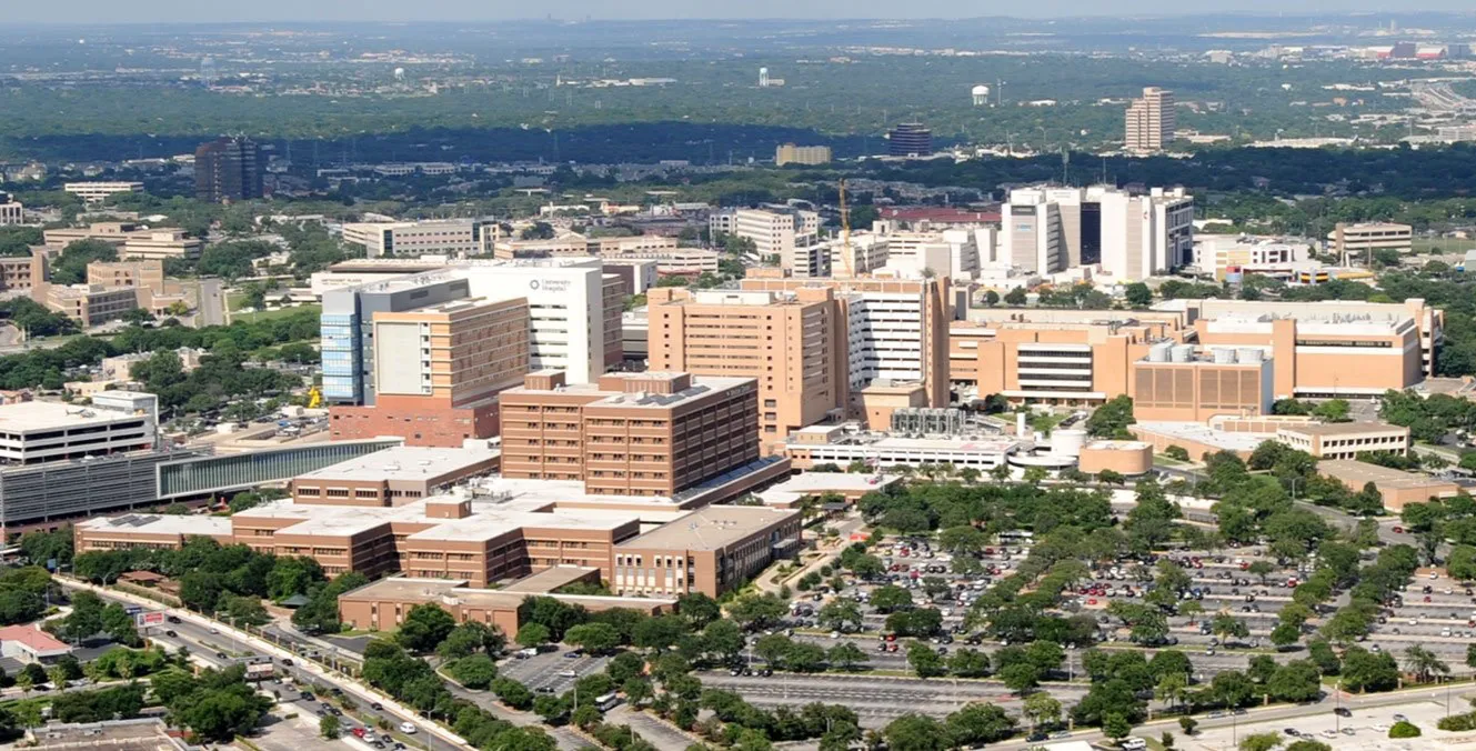 Aerial view of a large hospital complex with multiple beige and white buildings surrounded by trees and parking lots.