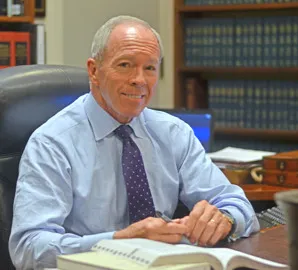 Middle-aged man in a blue shirt and purple tie sitting at an office desk with bookshelves in the background.