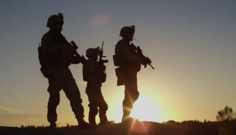 Silhouettes of three armed soldiers standing on a hill at sunset with clear sky.