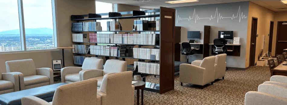 Professional office lounge with beige armchairs, bookshelves, computer stations, and a city view window.