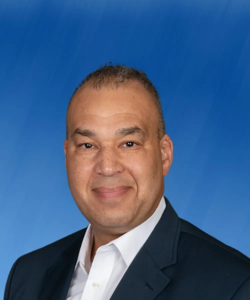 Professional headshot of a middle-aged man in a dark suit and white shirt against a blue background.