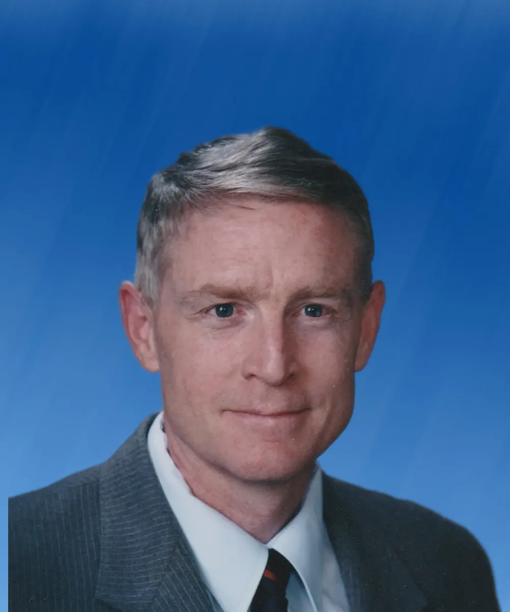 Portrait of a middle-aged man with short gray hair wearing a gray suit and tie against a blue background.