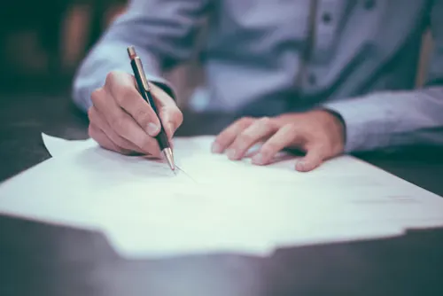 Person in blue shirt signing white documents with a black and silver pen.