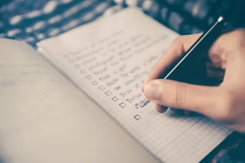 Close-up of a hand writing a checklist on graph paper with a black pen.