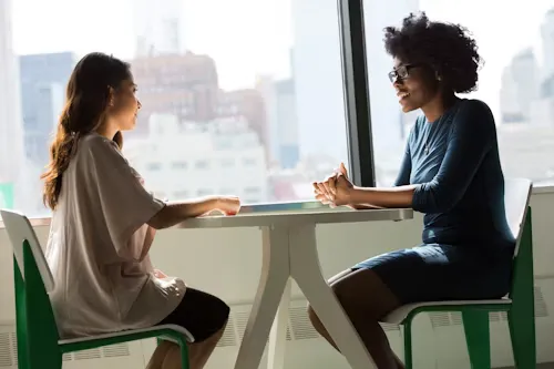 Two professional women engaged in conversation at a white table by a large window.